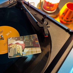 May include: A blue record player with a black vinyl record playing. A decorative coaster with a bedroom scene is on the record. A lit candle and a jack-o'-lantern decoration are in the background.