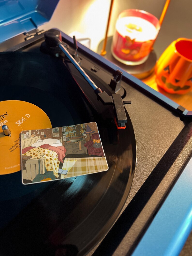 May include: A blue record player with a black vinyl record playing. A decorative coaster with a bedroom scene is on the record. A lit candle and a jack-o'-lantern decoration are in the background.