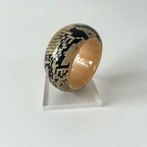 Puede incluir: Un brazalete ancho y redondo con un estampado de piel de serpiente en negro, beige y crema. El interior del brazalete es de color beige liso. El brazalete se exhibe sobre un soporte de acrílico transparente.