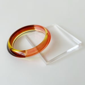 Könnte beinhalten: Ein durchscheinendes Armreif-Armband mit einem Farbverlauf von Dunkelbraun über Gelb zu Orange. Das Armband liegt auf einem transparenten, quadratischen Acrylblock. Schmuck.