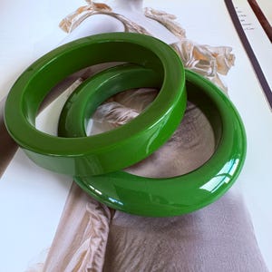 Könnte beinhalten: Zwei glänzende, grüne Armreifen. Die Armreifen sind kreisförmig und haben eine glatte Oberfläche. Sie scheinen aus einem festen Material zu bestehen. Der Schmuck wird vor einem neutralen Hintergrund präsentiert. Die Armreifen sind in einem leuchtenden Grünton gehalten.