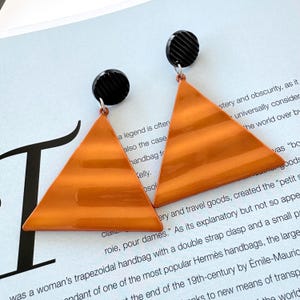 May include: A pair of orange triangle earrings with black circular tops. The earrings have a glossy finish and horizontal stripes on the triangle. The earrings are on a white surface with text.