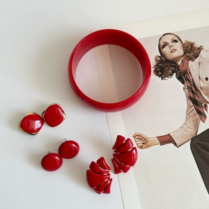 May include: A collection of vintage red jewelry, including a wide bangle bracelet and three pairs of earrings. The earrings feature gold accents and various designs, including a square, oval, and a segmented fan shape. The jewelry is displayed on a white surface.