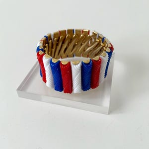 May include: A patriotic bracelet featuring alternating red, white, and blue segments. The bracelet has a gold-colored interior and is displayed on a clear acrylic stand. This stretch bracelet is a stylish accessory.