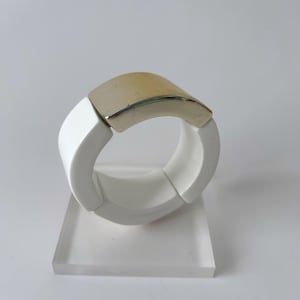May include: A white and gold bracelet with a curved, rectangular gold accent. The bracelet is made of white, curved segments. The jewelry is displayed on a clear, rectangular stand.