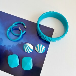 Puede incluir: Una colección de joyas turquesas, que incluye pendientes de aro, una pulsera de brazalete y varios pendientes de botón. Los pendientes incluyen tachuelas cuadradas y redondas con rayas blancas y azules. La pulsera tiene un diseño texturizado.