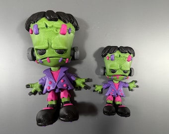 May include: Two green Frankenstein monster figurines. The figures have black hair, gray ear bolts, and are wearing purple jackets over pink shirts. The monsters have pink and purple stitches and black shoes. One figure is larger than the other.
