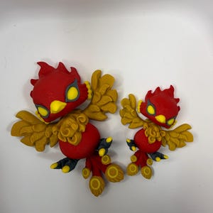 May include: Two red and gold bird figurines with yellow accents. The birds have detailed wings, feet, and head crests. The figurines are likely decorative items or toys.
