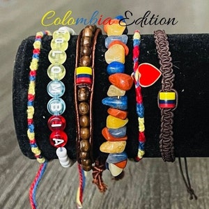 May include: Set of six colourful bracelets featuring a beaded bracelet with the word "COLOMBIA" spelled out, a wooden bead bracelet, a bracelet with colourful stones, a red and yellow string bracelet, a blue and yellow string bracelet, and a brown braided bracelet with a red heart charm and a Colombian flag charm. The bracelets are displayed on a black surface.