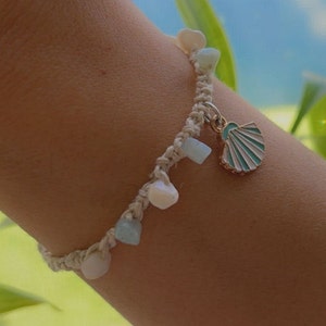 May include: A handmade macrame bracelet featuring small, light-colored gemstones and a seashell charm. The bracelet is adorned with white, blue, and cream-colored beads. The seashell charm is gold-toned with a turquoise and white enamel design.