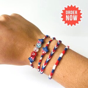 May include: A set of four bracelets featuring red, white, and blue colours. One bracelet includes star-shaped beads and round beads spelling USA. The other three bracelets are made of braided thread in patriotic patterns of red, white, and blue stripes.