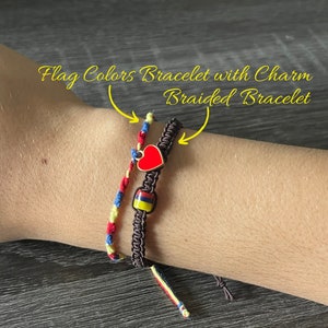 May include: Two bracelets on a wrist. One is a braided brown bracelet with a red heart charm and a small bead with the colours of the Colombian flag. The other is a string bracelet with red, yellow, and blue strings.