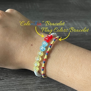 May include: Two bracelets on a person's wrist. One bracelet is made of white beads with the word "COLOMBIA" spelled out in black letters. The other bracelet is made of yellow, red, and blue string.