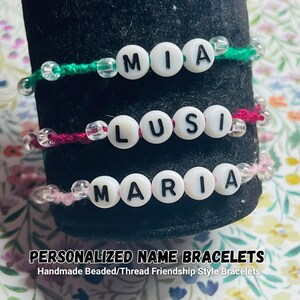 May include: Three bracelets made of green, dark pink, and light pink thread, each featuring white round beads with black letters spelling out the names MIA, LUSI, and MARIA, accented with clear transparent beads on the ends of each name segment.