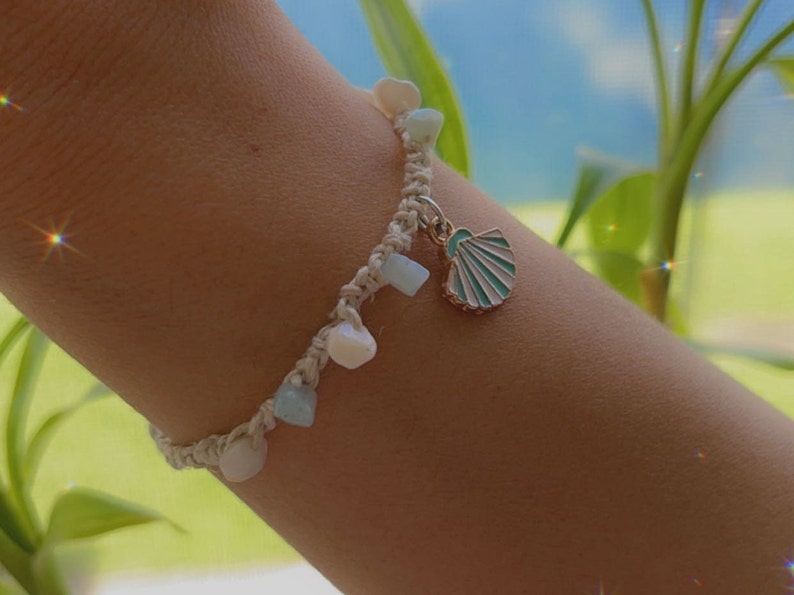 May include: A handmade macrame bracelet featuring small, light-colored gemstones and a seashell charm. The bracelet is adorned with white, blue, and cream-colored beads. The seashell charm is gold-toned with a turquoise and white enamel design.