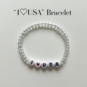 May include: A circular beaded bracelet featuring clear, translucent beads and four white round beads with black lettering and a red heart symbol that spell out I heart USA. The text I heart USA Bracelet is printed in black above the jewellery item.