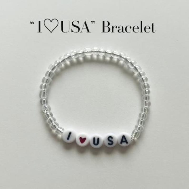 May include: A circular beaded bracelet featuring clear, translucent beads and four white round beads with black lettering and a red heart symbol that spell out I heart USA. The text I heart USA Bracelet is printed in black above the jewellery item.