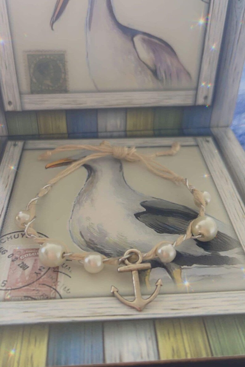 May include: A nautical-themed bracelet with faux pearls and a silver anchor charm. The bracelet is displayed in a decorative box with a seagull illustration and a twine bow.
