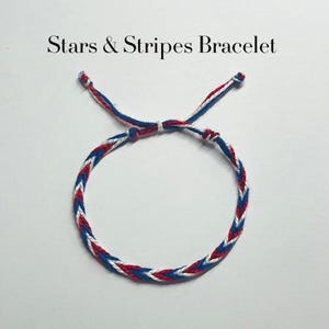 May include: A braided bracelet featuring red, white, and blue thread in a chevron pattern with adjustable tie ends. The text Stars & Stripes Bracelet is printed above the item. The design consists of alternating coloured strands woven together.