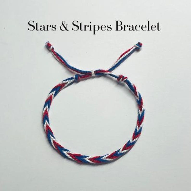May include: A braided bracelet featuring red, white, and blue thread in a chevron pattern with adjustable tie ends. The text Stars & Stripes Bracelet is printed above the item. The design consists of alternating coloured strands woven together.
