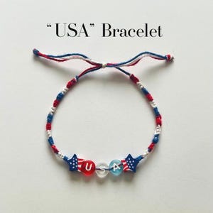 May include: A bracelet featuring red, white, and blue braided cord with two star-shaped beads patterned like the American flag, flanking three round beads spelling out USA in white letters. The text USA Bracelet is printed above the item.