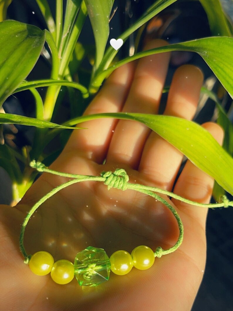Puede incluir: Un brazalete de cuerda verde con cuatro cuentas verdes y una cuenta verde facetada en el centro.