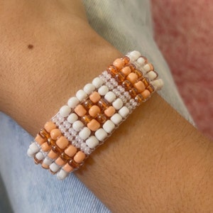 K&ouml;nnte beinhalten: Ein Perlenarmband mit einem gewebten Muster aus wei&szlig;en, pfirsichfarbenen und bernsteinfarbenen Perlen. Das Armband wird am Handgelenk getragen. Die Perlen sind klein und quadratisch und erzeugen eine strukturierte Oberfl&auml;che. Das Armband ist wahrscheinlich handgefertigt.
