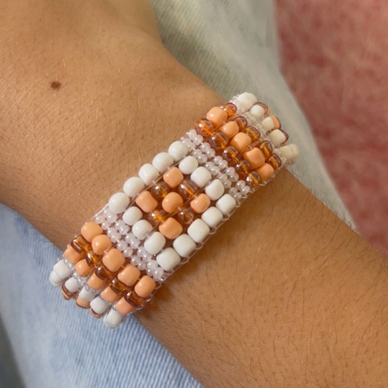 K&ouml;nnte beinhalten: Ein Perlenarmband mit einem gewebten Muster aus wei&szlig;en, pfirsichfarbenen und bernsteinfarbenen Perlen. Das Armband wird am Handgelenk getragen. Die Perlen sind klein und quadratisch und erzeugen eine strukturierte Oberfl&auml;che. Das Armband ist wahrscheinlich handgefertigt.