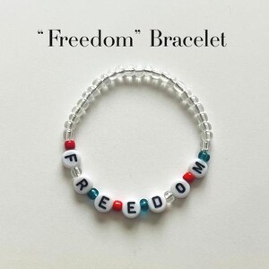 May include: A circular beaded bracelet featuring the word Freedom spelt out with white alphabet beads with black lettering. The design includes clear, red, and teal accent beads interspersed throughout the strand. The text Freedom Bracelet is visible.