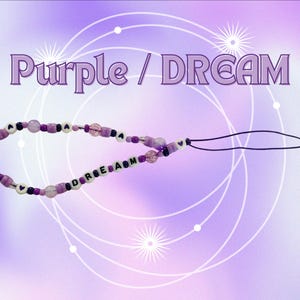 May include: A phone charm featuring a variety of purple, lavender, and clear beads, including white letter beads spelling out the word DREAM and small heart-shaped beads. The charm is attached to a thin black cord loop for easy attachment to devices.