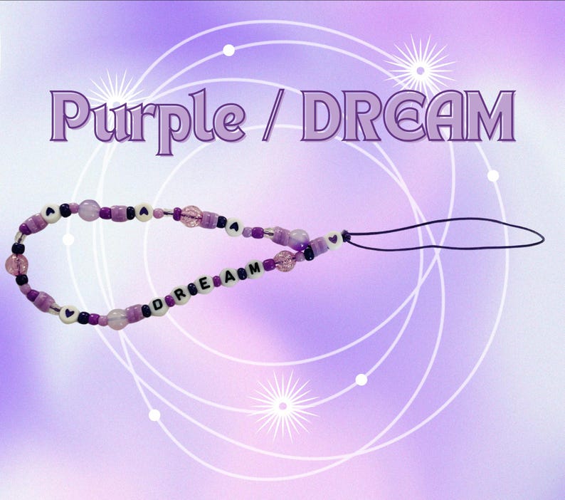 May include: A phone charm featuring a variety of purple, lavender, and clear beads, including white letter beads spelling out the word DREAM and small heart-shaped beads. The charm is attached to a thin black cord loop for easy attachment to devices.