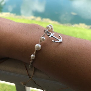 May include: A handmade bracelet featuring faux pearls and a silver anchor charm. The bracelet is made with a natural-colored cord and is designed to be worn on the wrist. The background shows a body of water and green foliage.
