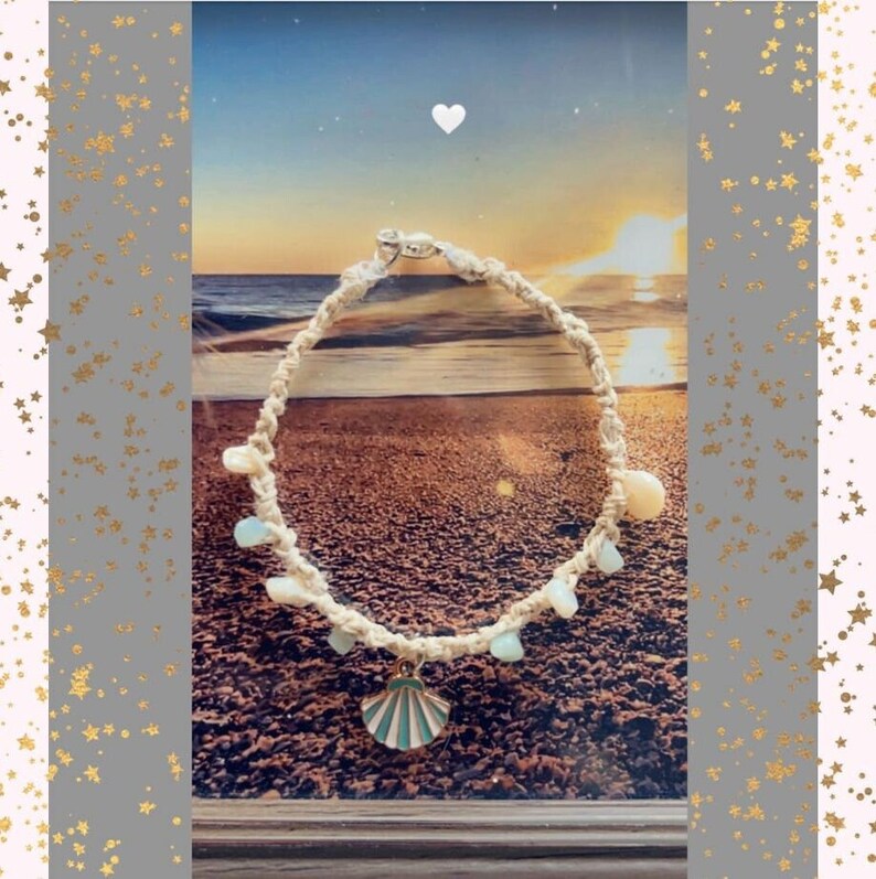 May include: A cream-colored macrame anklet with a seashell charm and light blue and white beads. The charm is a seashell with a teal and white design. The anklet is displayed against a beach scene.