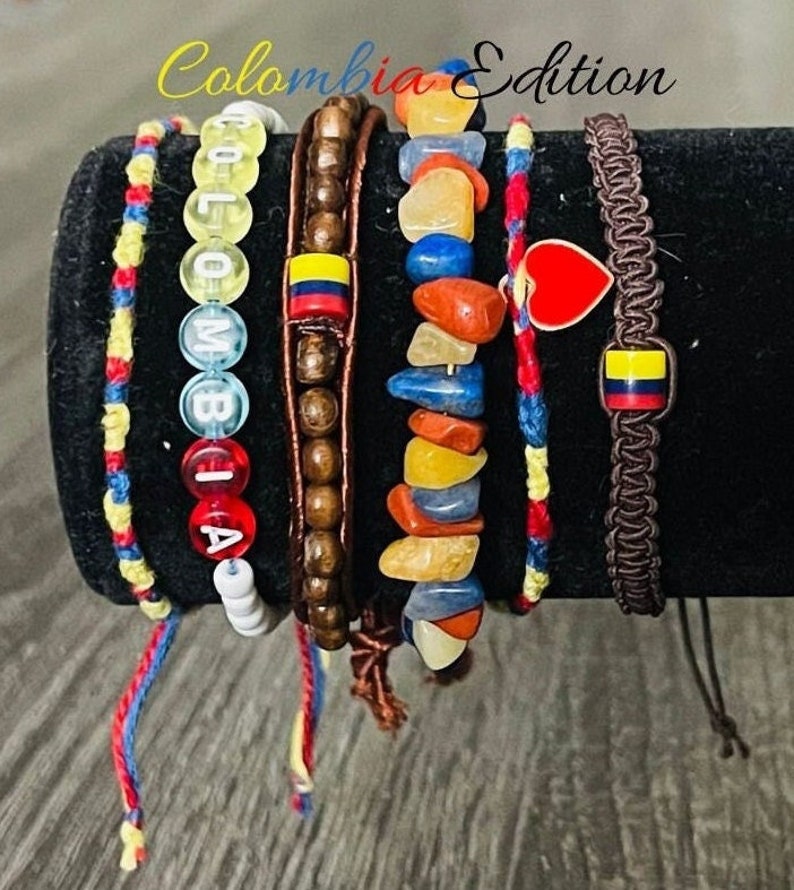 May include: Set of six colourful bracelets featuring a beaded bracelet with the word "COLOMBIA" spelled out, a wooden bead bracelet, a bracelet with colourful stones, a red and yellow string bracelet, a blue and yellow string bracelet, and a brown braided bracelet with a red heart charm and a Colombian flag charm. The bracelets are displayed on a black surface.