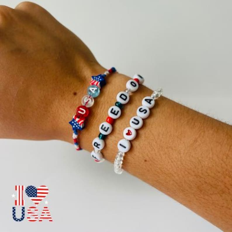 May include: Three beaded bracelets featuring red, white, and blue colour schemes. One bracelet displays the text USA with star-shaped beads. Another spells FREEDOM with round white beads. The third bracelet features the text I heart USA.