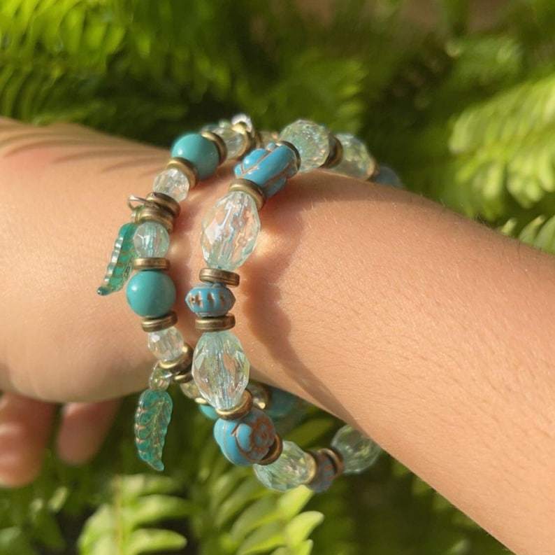 Puede incluir: Dos pulseras de cuentas de vidrio turquesa y transparentes con detalles dorados y abalorios en forma de hoja.