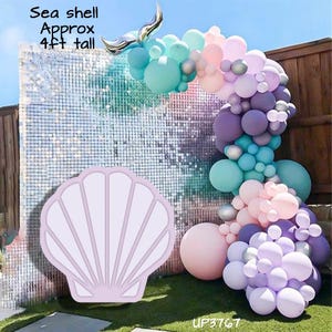 May include: A balloon arch featuring shades of teal, purple, pink, and silver, accented by a silver foil seashell topper. A large, light purple seashell cutout is displayed in the foreground, with text stating Sea shell Approx 4ft tall.