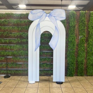 May include: A white archway with a large blue bow on top. The archway is made of cardboard and is standing on a tiled floor.