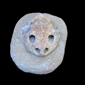 Puede incluir: Una escultura de piedra tallada de una cara con dos agujeros para los ojos y dos para las fosas nasales, colocada sobre una piedra más grande, de forma ovalada. La escultura es de color beige y marrón, con una textura rugosa, sobre un fondo negro.
