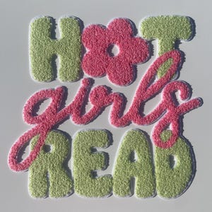 May include: A green and pink felt patch with the text "Hot girls read" in a retro style font. The word "girls" is in pink and the rest of the text is in green. There is a pink flower in the middle of the patch.