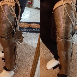 Pode incluir: Um close-up de um par de armaduras de perna de metal prateadas com uma barbatana decorativa no lado. A armadura é usada na perna de uma pessoa e é presa com correias.