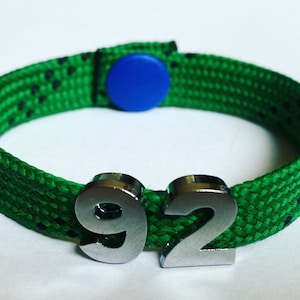 May include: Green and black braided bracelet with a blue button closure and silver metal number charms that read "92".