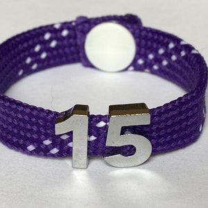 Peut inclure: Bracelet tressé violet et blanc avec des breloques en métal argenté numérotées qui épellent "15".