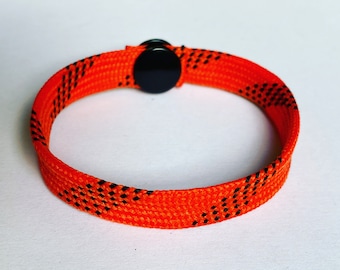 May include: Orange bracelet with black accents. The bracelet is made of woven material and features a black circular clasp. The design includes small black dashes throughout the orange band. A simple, yet stylish accessory.