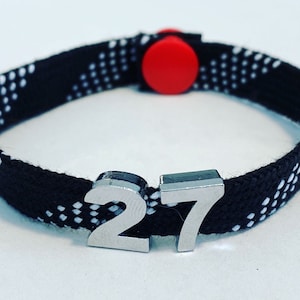 May include: Black and white patterned bracelet with a red adjustable clasp and silver metal number charms that read "27".