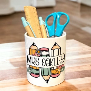 May include: White ceramic pencil holder featuring a design of three pencils with flowers and a banner that reads "Mrs. Oakley". The holder contains two blue pens, a wooden ruler, and blue scissors.