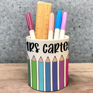 May include: White ceramic pencil holder with a colourful pencil design and the text "Mrs Carter" on the front. The holder is filled with pens and a ruler.