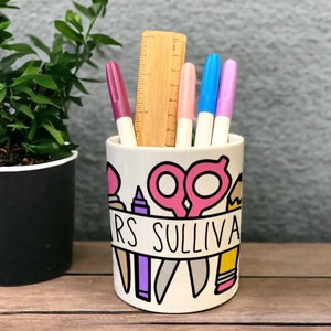 May include: White ceramic pencil holder with a colourful design of school supplies, including scissors, a pencil, a ruler, and a paintbrush. The holder has the text "Mrs. Sullivan" on a banner.