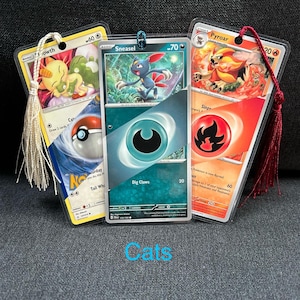 May include: Three Pokémon card bookmarks. The cards are encased in clear plastic and have tassels. The cards feature illustrations of Pokémon characters. The word "Cats" is written in blue cursive at the bottom.