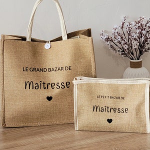 May include: A tan jute tote bag and a matching rectangular pouch, both featuring white trim and black text that reads LE GRAND BAZAR DE Ma&icirc;tresse and LE PETIT BAZAR DE Ma&icirc;tresse, each accented with a small black heart icon and a white button closure.